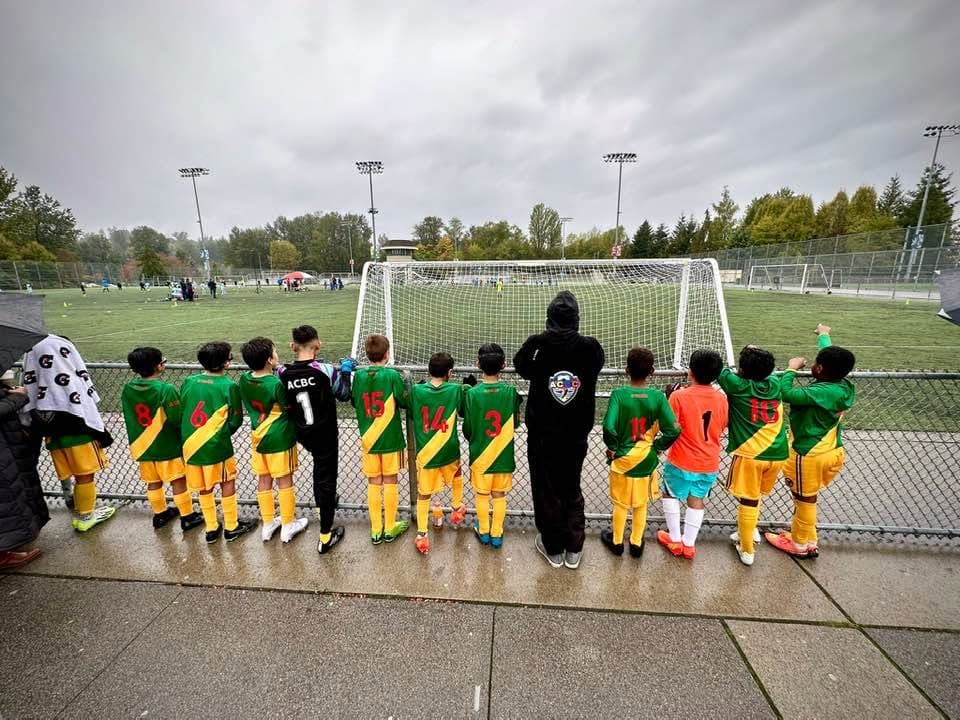 ACBC players in green and gold along the touchline at a rainy Burnaby pitch